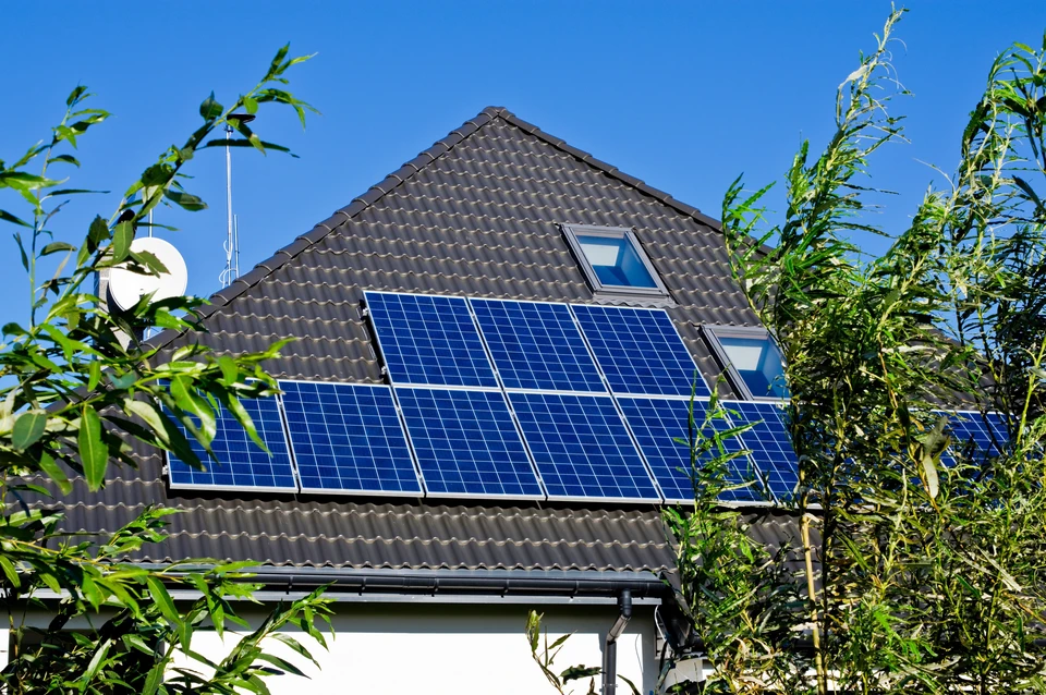 Maison résidentielle avec toiture en tuiles grises équipée de panneaux solaires photovoltaïques bleus, lucarne avec fenêtre de toit, antenne parabolique blanche, végétation luxuriante au premier plan par temps ensoleillé