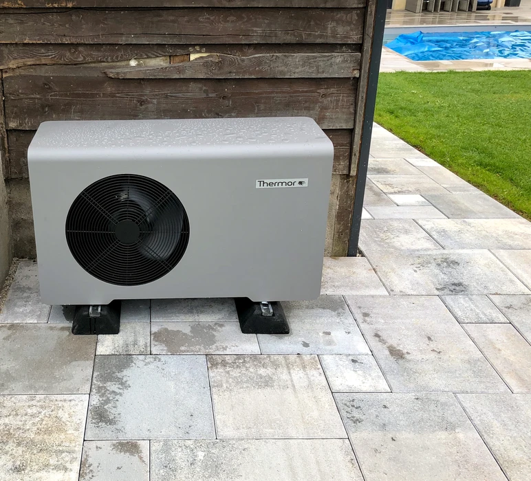 Unité extérieure de pompe à chaleur air/eau Thermor, installée sur plots antivibratoires noirs, posée sur une terrasse en dalles de béton