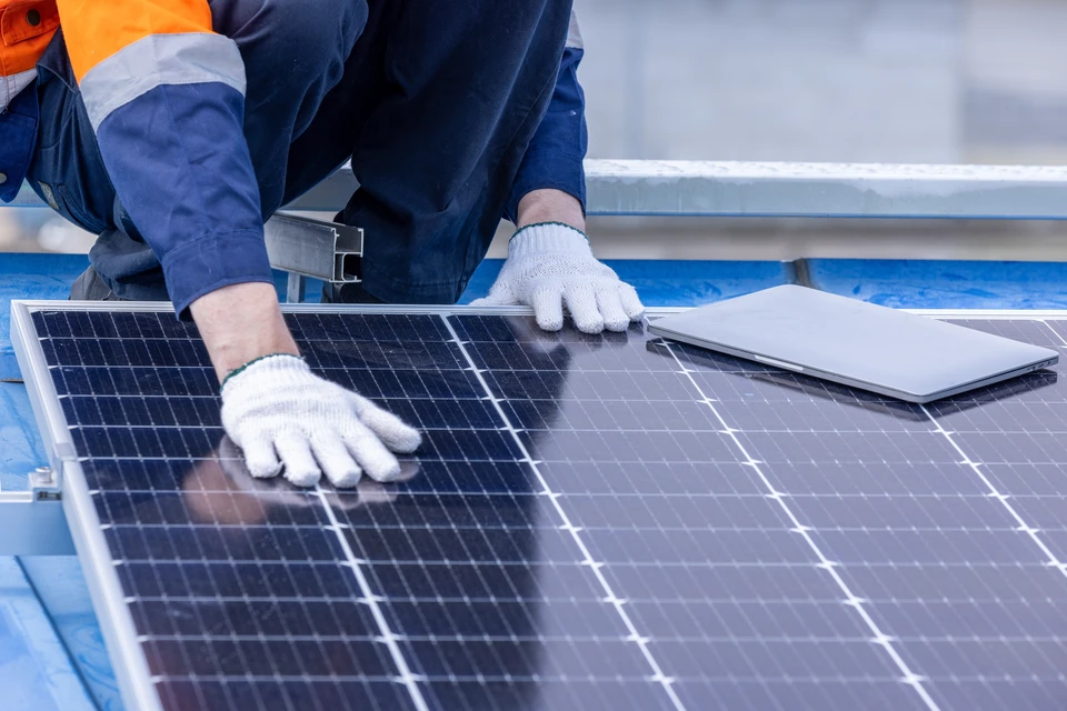 Deux techniciens en combinaisons vertes et casques blancs installent des panneaux photovoltaïques sur une toiture résidentielle en tuiles sous un ciel bleu dégagé, environnement verdoyant visible en arrière-plan